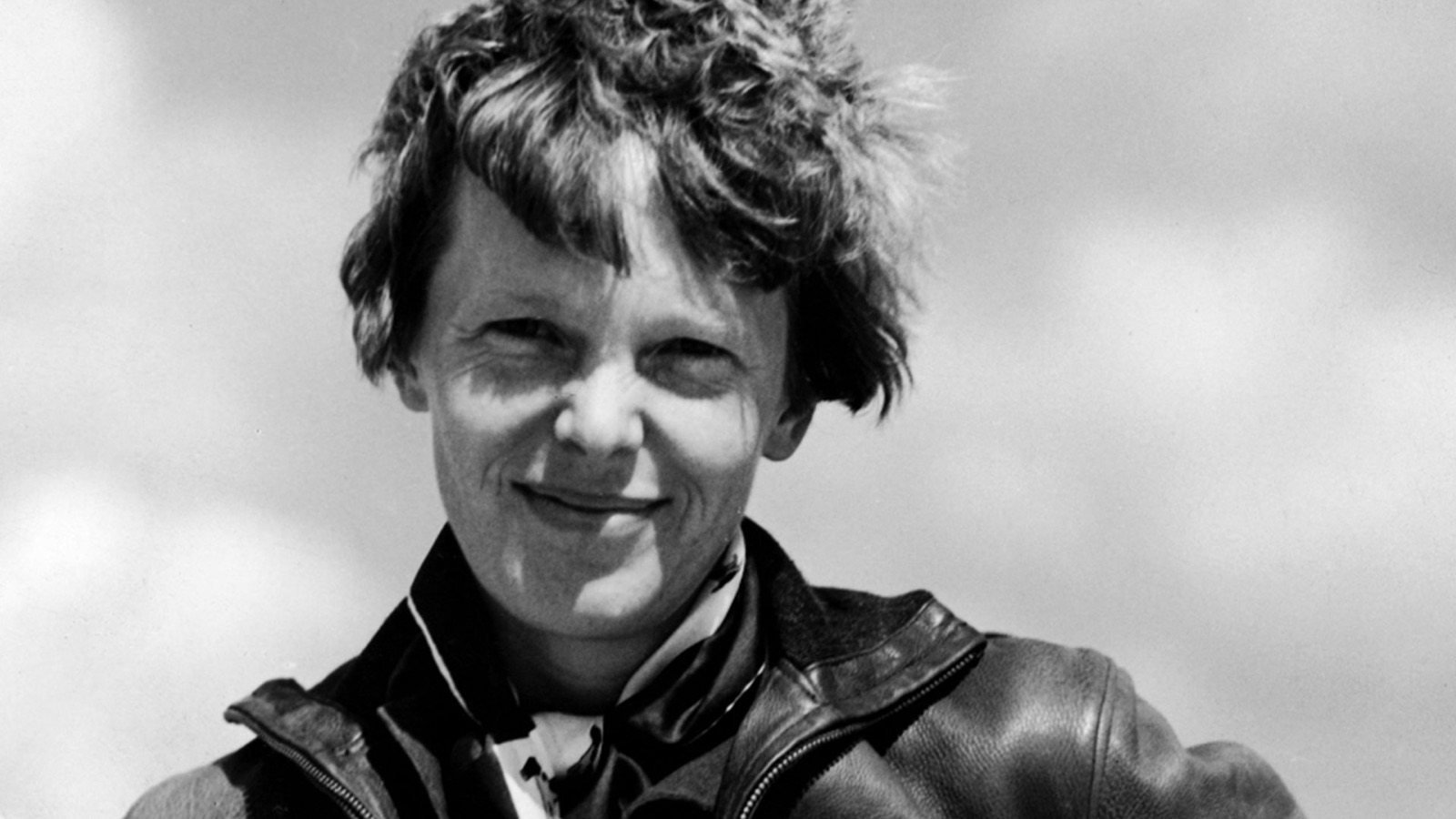 Amelia Earhart, smiling, in a leather flying jacket against a cloudy sky